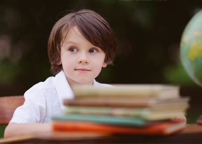 a little boy sitting at a desk with a stack of books on the desk