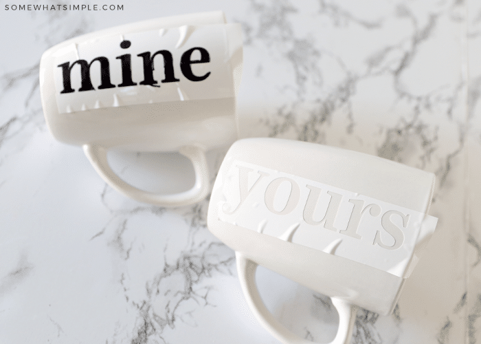 two white mugs on the counter, one with black letters that spell out "mine" and the other with a whit stencil attached to it that says "yours"